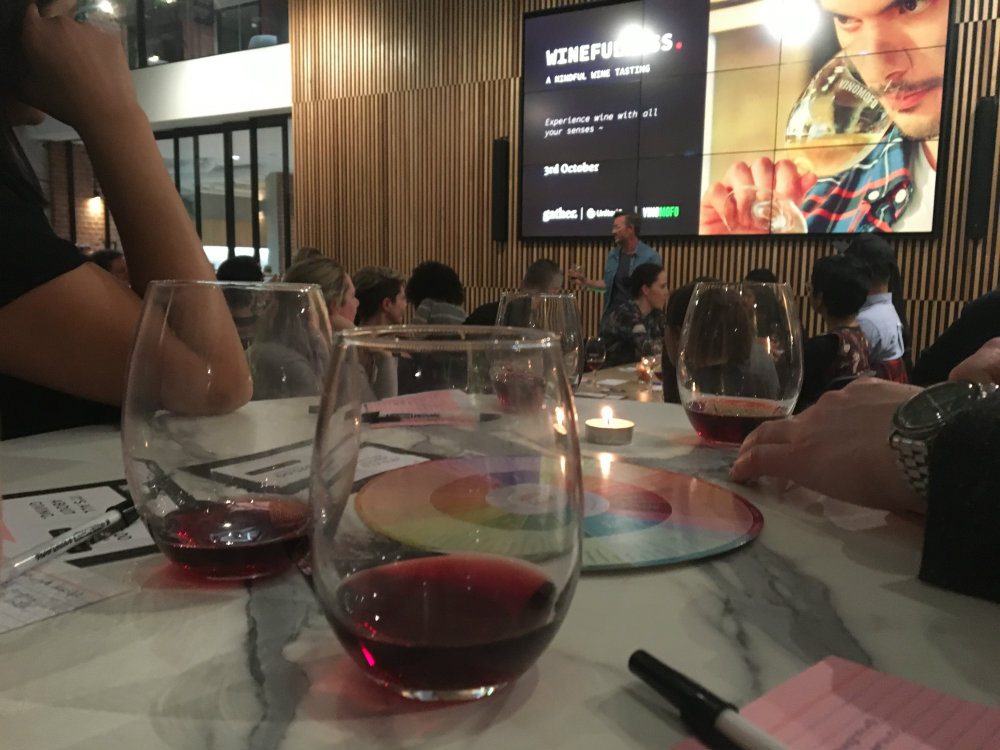 glasses of red wine at a table with prompt cards, pens and a person speaking at the front of the room with a projected image of a man tasting wine behind him