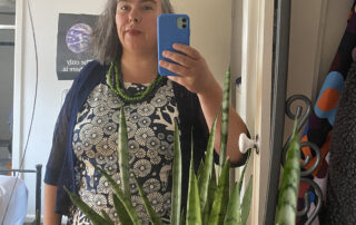 a photo of me holding my phone in front of a mirror with a plant and a patterned top. I have a strange kind of expression on my face. It is kind of a 'not sure what that was all about' kind of expression